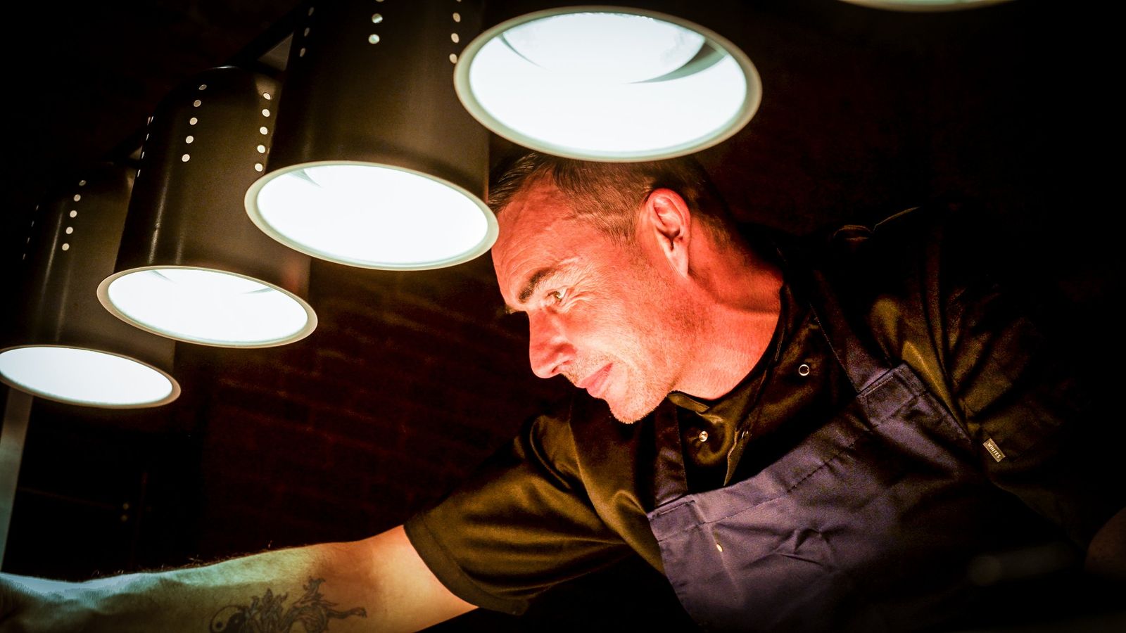 A chef leaning in to finish a dish under the warm pass lights