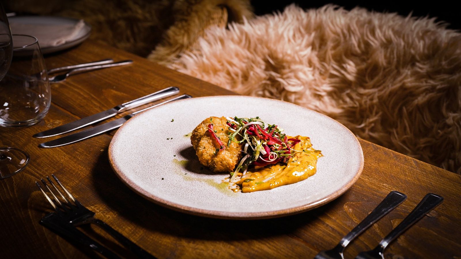 A plated course of a golden croquette with dressed leaves and sauce, on a wooden table laid for dinner